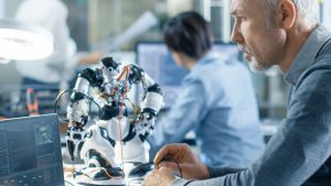 Senior Electronics Engineer Works With Robot, Programming it with Laptop. In the Background Laboratory with Technical Personnel Working.